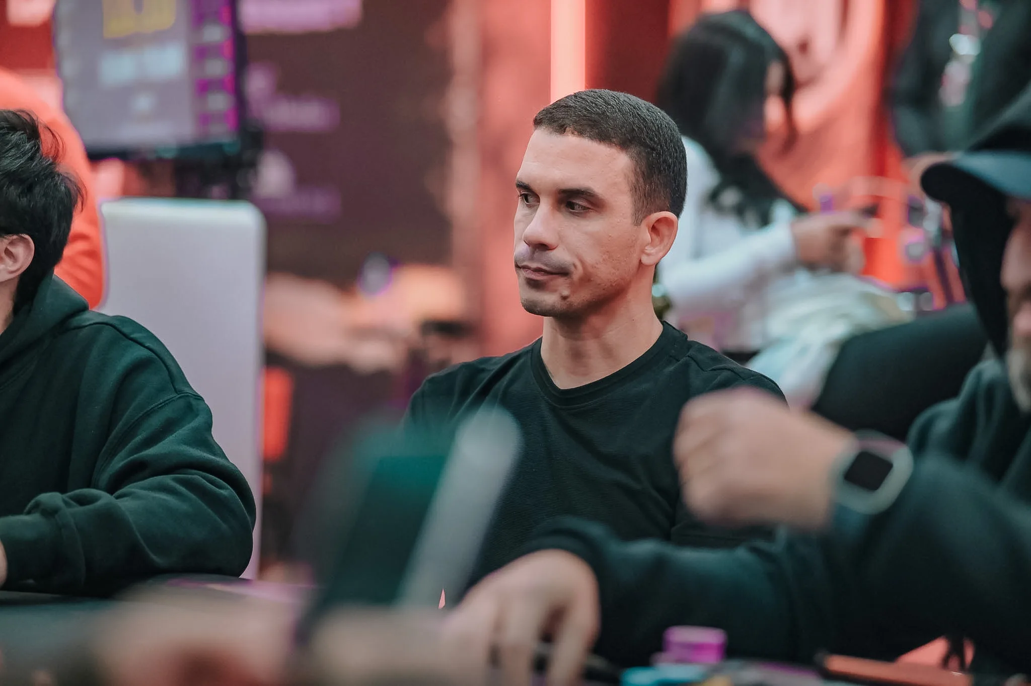 A poker player looking intently at their cards during a high-stakes tournament, with other players and chips visible on the table.
